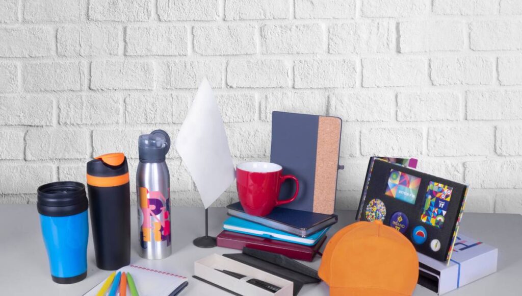 A variety of promotional products, like journals and cups, arranged with ample copy space against a grey wall background