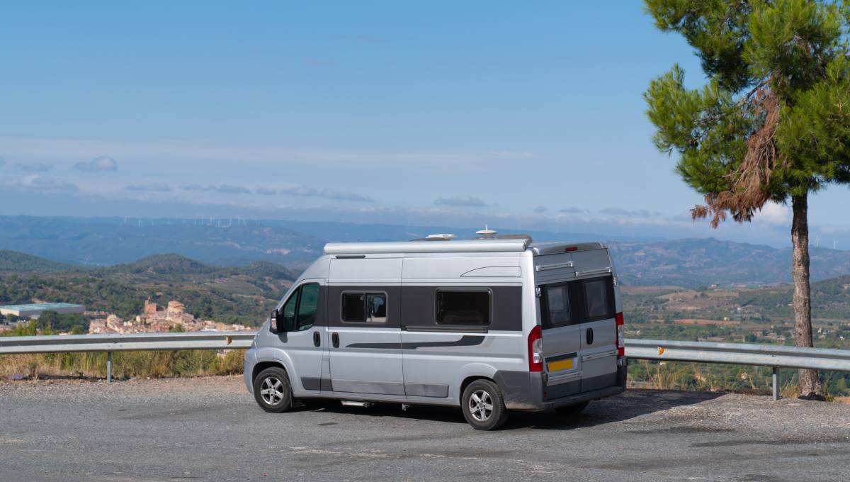 A silver conversion van is parked near a scenic overlook in Spain. In the distance, a town, mountains, and fields are visible.