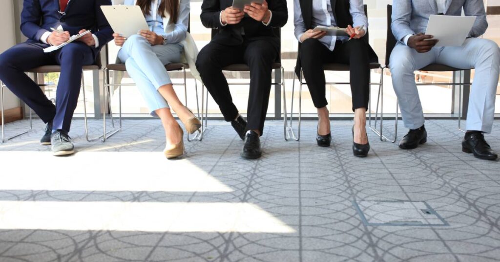 Three men and two women are sitting inside a building. They are wearing professional attire and filling out paperwork.