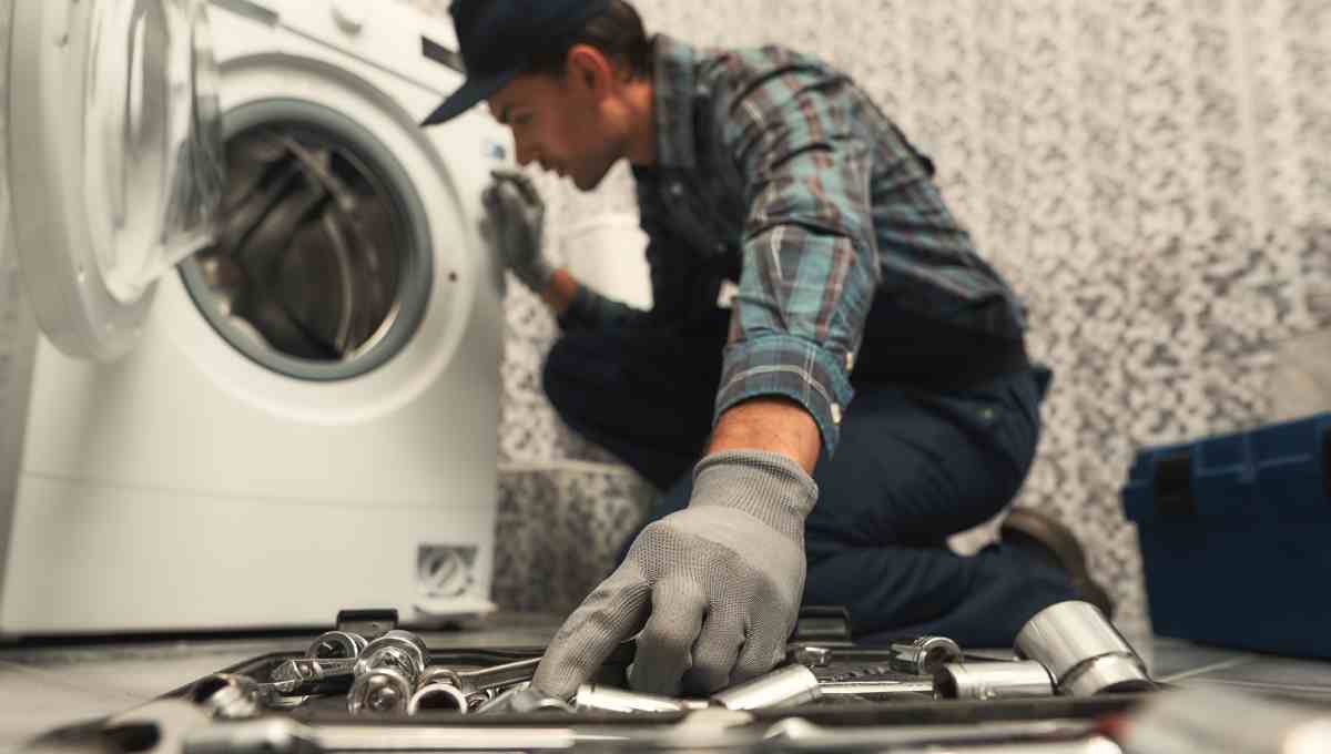 A plumber with gloves on repairs a washing machine using a wrench with various tools scattered around the floor.