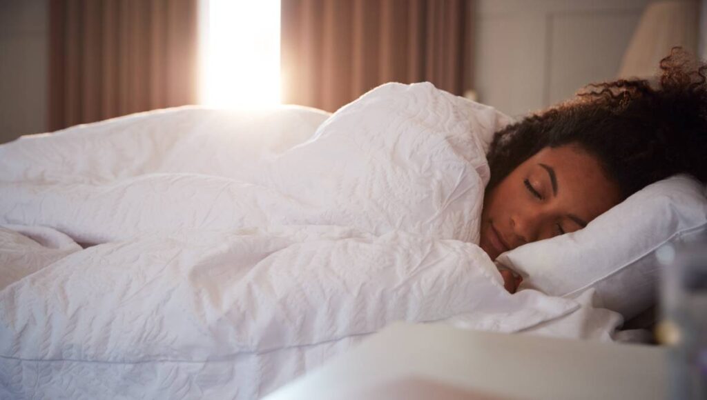 A woman lies in bed asleep under a fluffy white bedspread. The sun shines in behind her between two curtains.