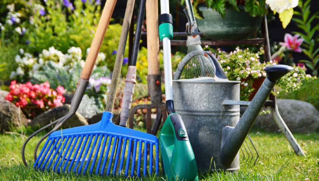 A wide selection of gardening and farming tools all lined up. Among them are a rake, pitchfork, and watering can.