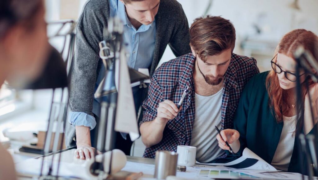 A group of young professionals collaborate on a project in a modern office setting using computers and documents.