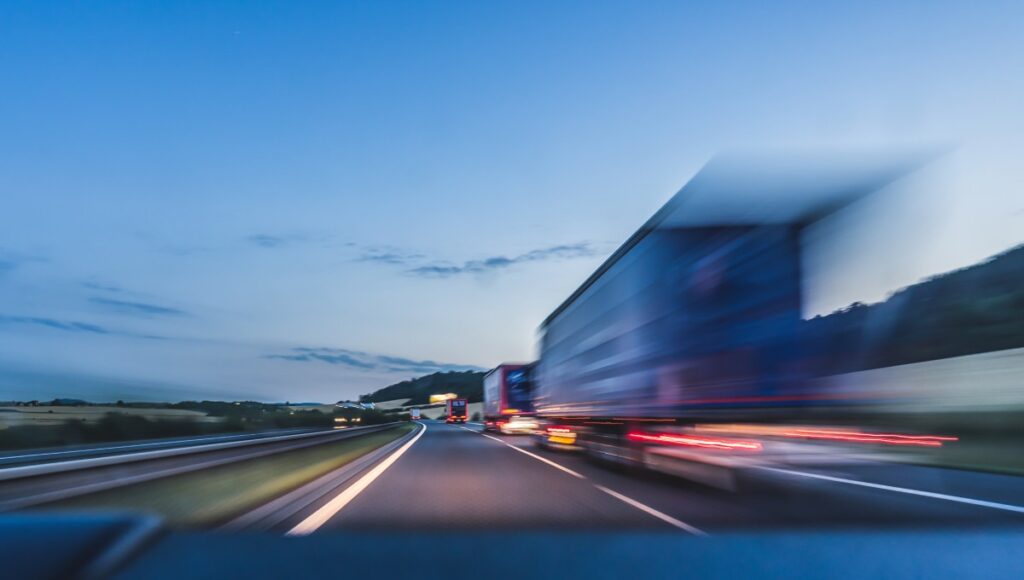 A blurry view shows a busy four-lane interstate at dusk. Many tractor-trailers are driving down the interstate.