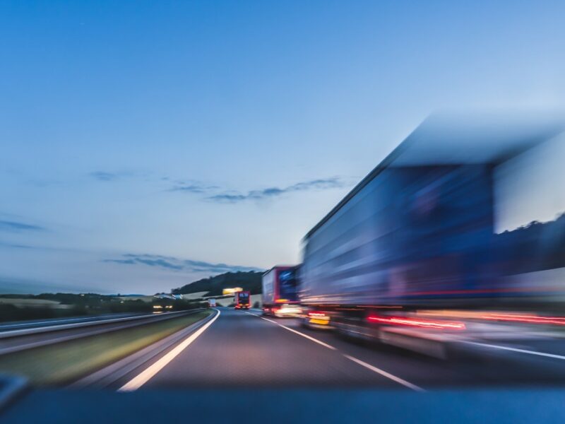 A blurry view shows a busy four-lane interstate at dusk. Many tractor-trailers are driving down the interstate.