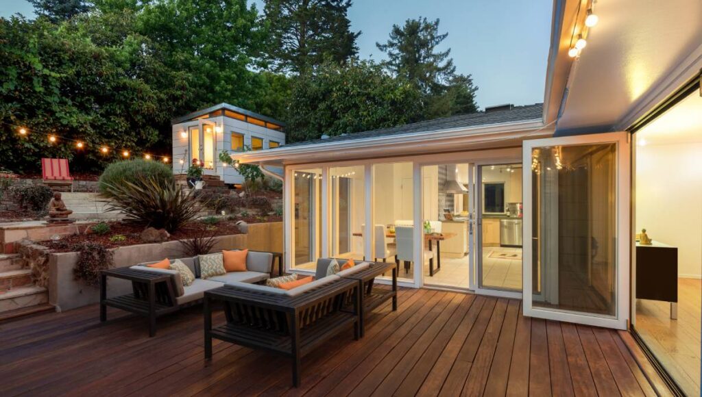 A dark wood deck is illuminated by string lights. There are two couches on the deck and raised landscaping behind them.