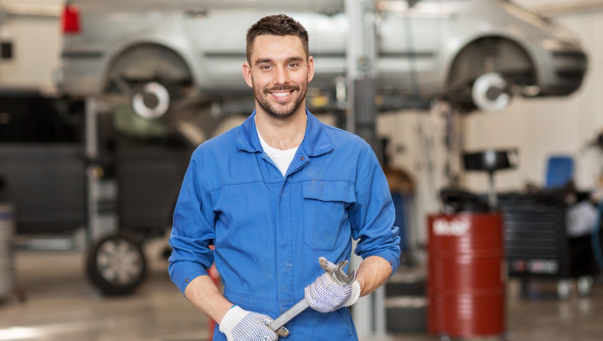 A man wearing a blue jumpsuit and holding a wrench. He stands in an auto garage with a minivan elevated in the background.