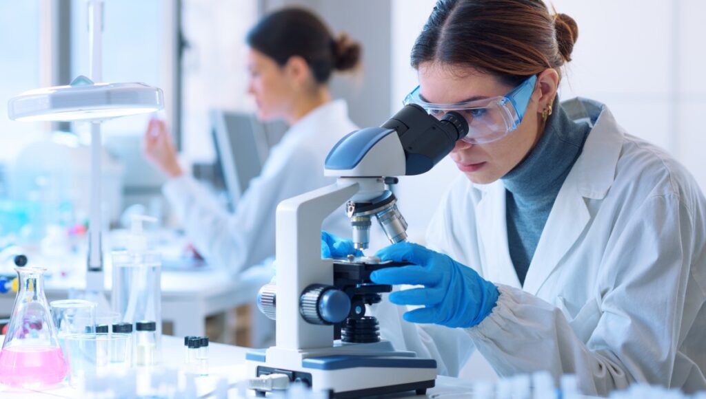 A female researcher wearing protective eye gear, blue gloves, and a lab coat looks through a white and blue microscope.