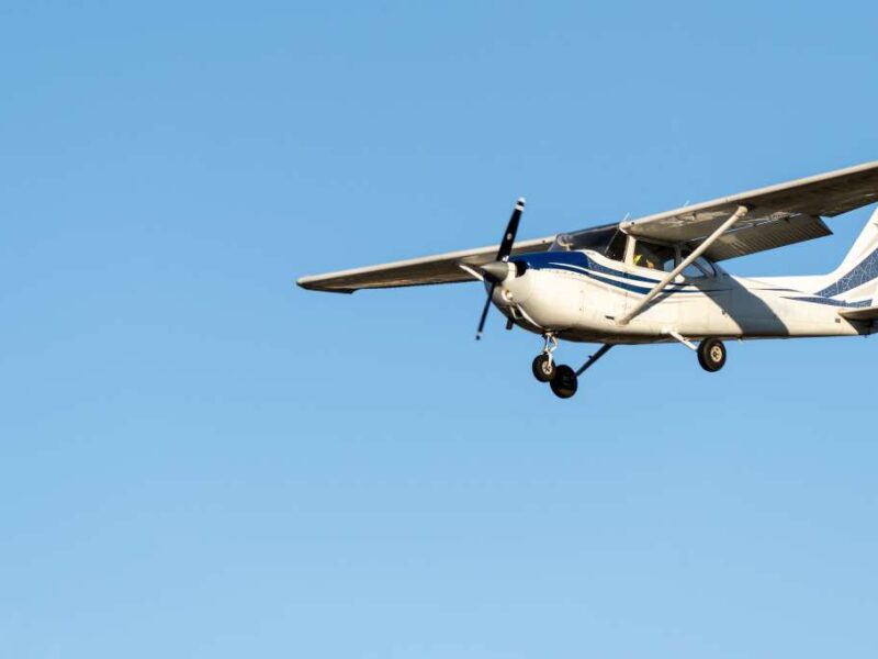 A small white airplane with blue lines on it flies in the sky on a sunny day. There are no clouds.