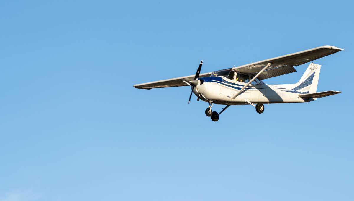 A small white airplane with blue lines on it flies in the sky on a sunny day. There are no clouds.