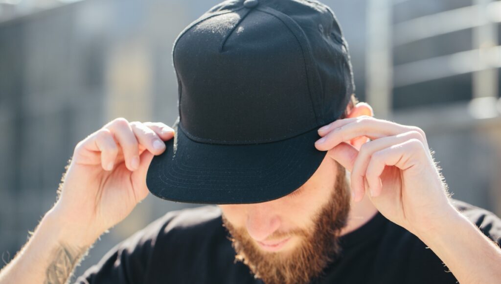 A bearded man with a forearm tattoo tilts his head down while touching the brim of his black baseball cap.