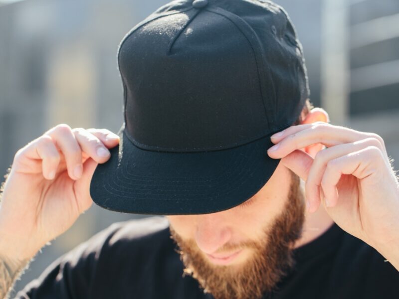 A bearded man with a forearm tattoo tilts his head down while touching the brim of his black baseball cap.