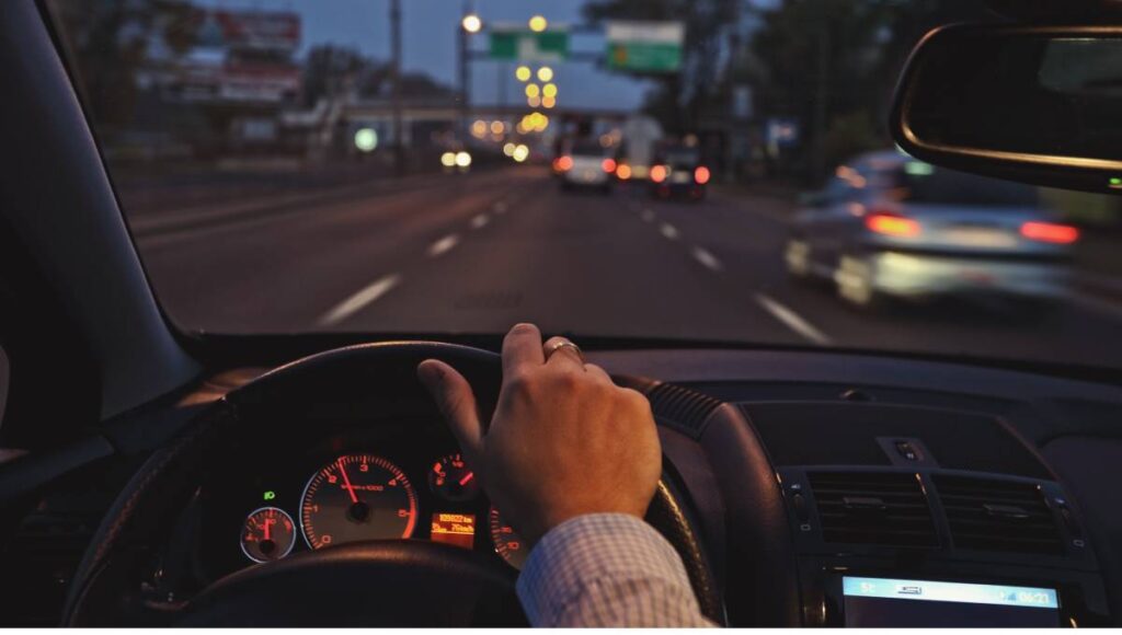 A person with their hand on the wheel drives their car down the highway with blurry cars and signs in the distance.