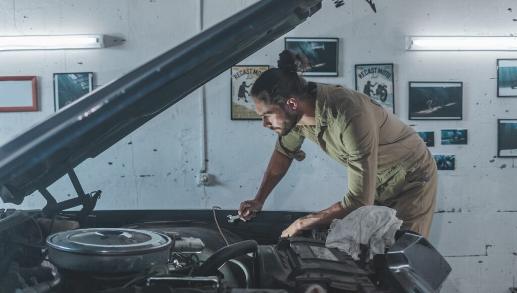 A mechanic in a beige uniform works on an old classic car with the engine compartment open in a garage.