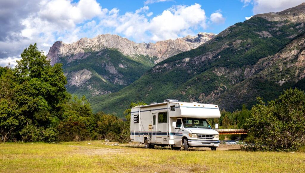 A panoramic view of a white RV parked in front of a large mountain range on a sunny day. There are trees surrounding the RV.