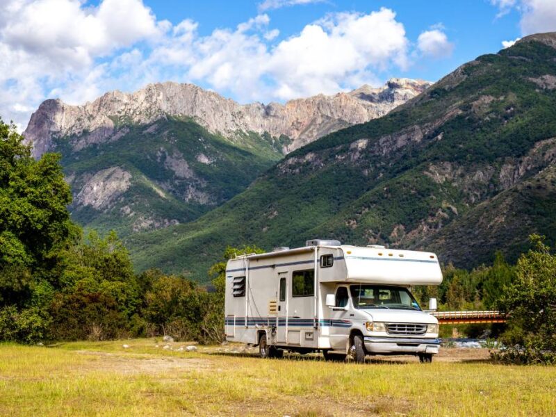 A panoramic view of a white RV parked in front of a large mountain range on a sunny day. There are trees surrounding the RV.