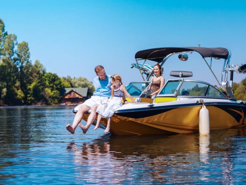 A father and daughter sit on the front part a stopped boat, dangling their feet above the water. A woman stands behind the wheel.