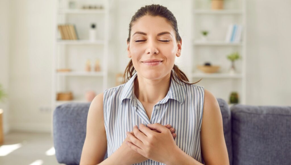 A woman sitting on a couch in a living room with her eyes closed, a soft smile, and her hands clasped gently over her chest.