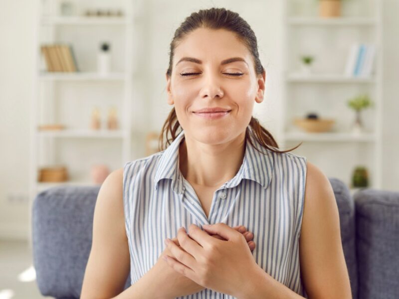 A woman sitting on a couch in a living room with her eyes closed, a soft smile, and her hands clasped gently over her chest.