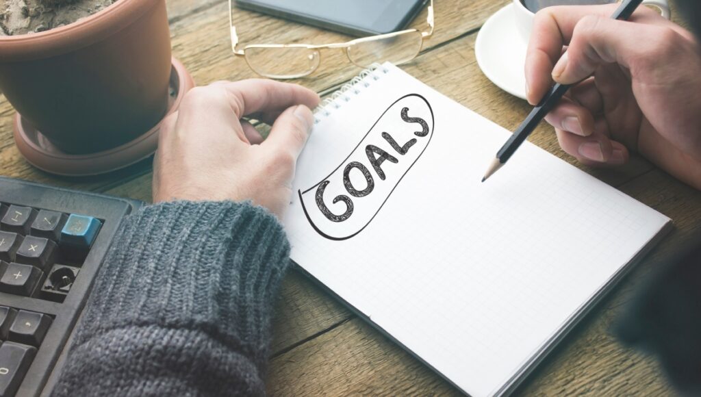 A man writes "goals" in a notebook with a black pencil. His glasses are above the notebook, and a coffee cup is to his right.