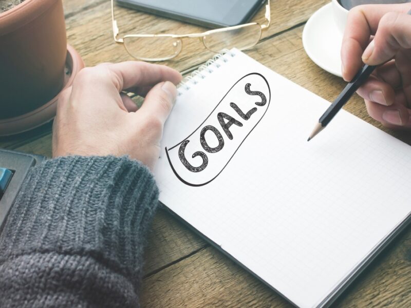 A man writes "goals" in a notebook with a black pencil. His glasses are above the notebook, and a coffee cup is to his right.