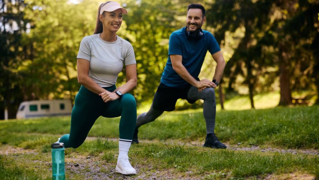 A man and woman in athletic gear smiling while doing lunges together outdoors. The woman has a water bottle nearby.