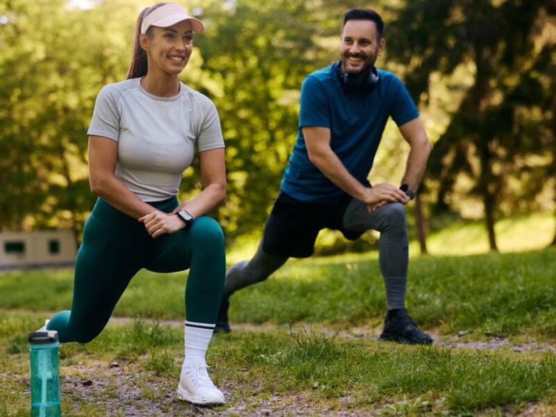 A man and woman in athletic gear smiling while doing lunges together outdoors. The woman has a water bottle nearby.