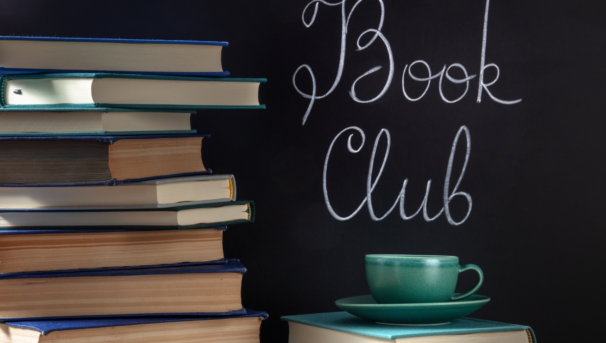 Two stacks of books, one with a teacup on top, in front of a black chalkboard with “book club” written on it.