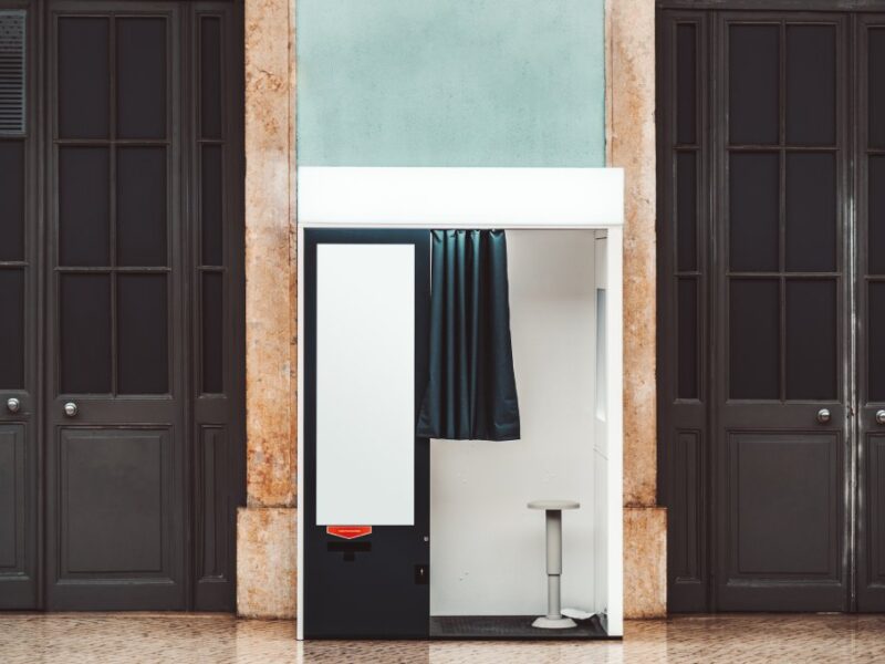 A black and white photo booth sits on a wooden floor and against a dark-wood wall. A black curtain on the booth is open.