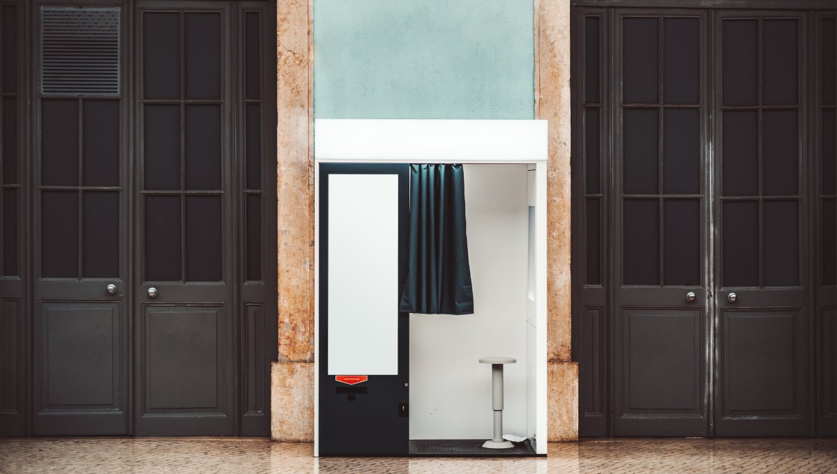 A black and white photo booth sits on a wooden floor and against a dark-wood wall. A black curtain on the booth is open.