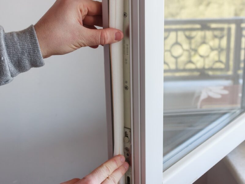 A close-up of a person applying foam insulation tape to the inside edge of an open casement window.