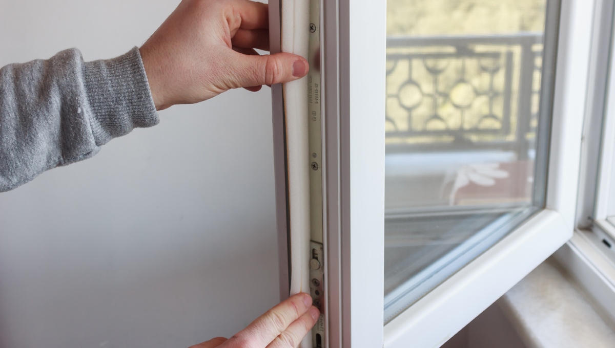 A close-up of a person applying foam insulation tape to the inside edge of an open casement window.