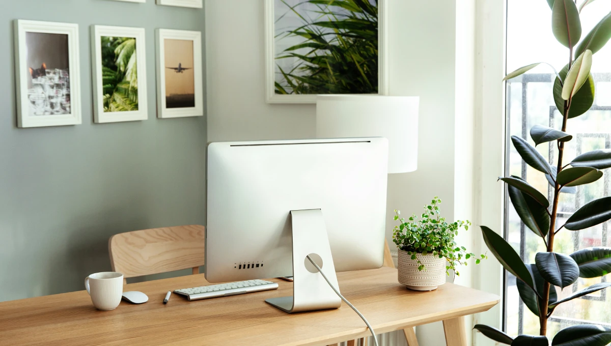 A home office with a computer, keyboard, and coffee mug on the desk. The walls have pictures, and a plant is in the corner.