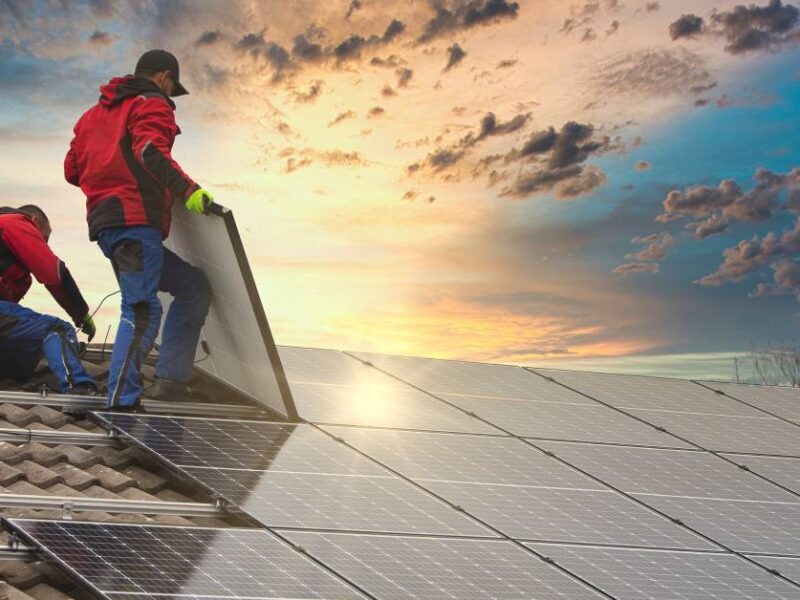 Two professional solar energy technicians in matching red jackets installing solar panels onto a residential home’s roof.