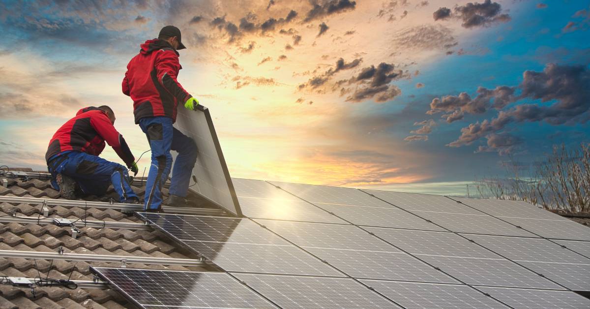 Two professional solar energy technicians in matching red jackets installing solar panels onto a residential home’s roof.