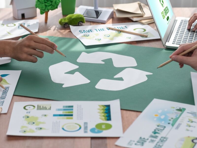 A table top is seen with several individuals seen from the neck down holding pencils over a recycling symbol.