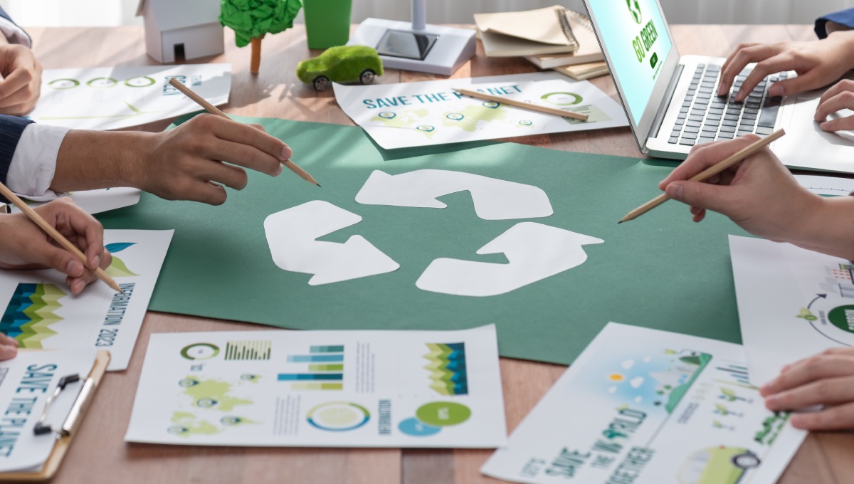 A table top is seen with several individuals seen from the neck down holding pencils over a recycling symbol.