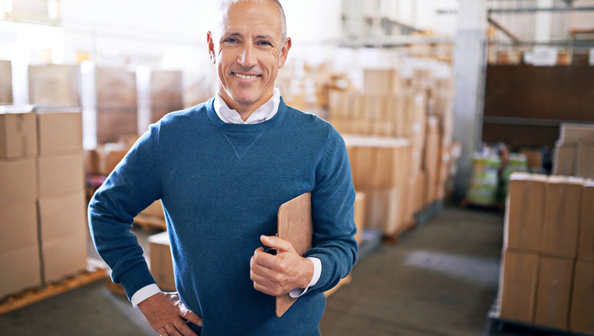 A man in a blue sweater standing in front of boxes stacked on pallets in a warehouse. He is smiling and holding a clipboard.