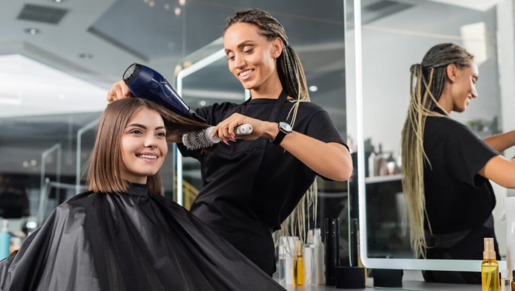 A salon professional brushes and dries the hair of a young woman while she sits in the styling chair.