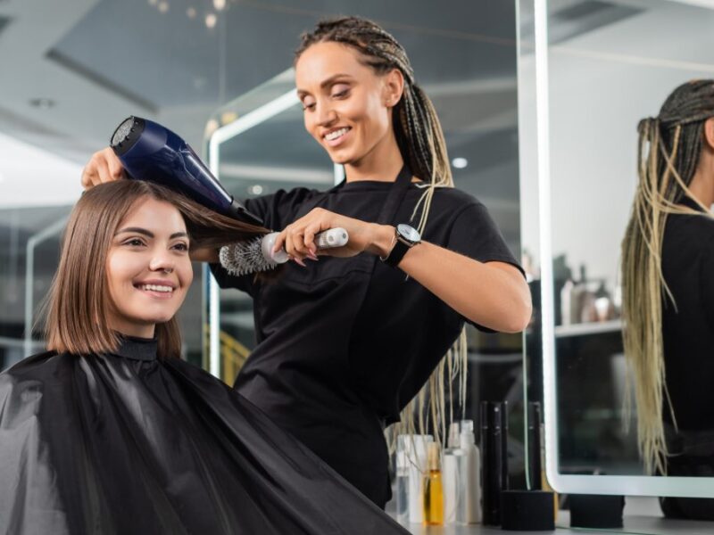 A salon professional brushes and dries the hair of a young woman while she sits in the styling chair.