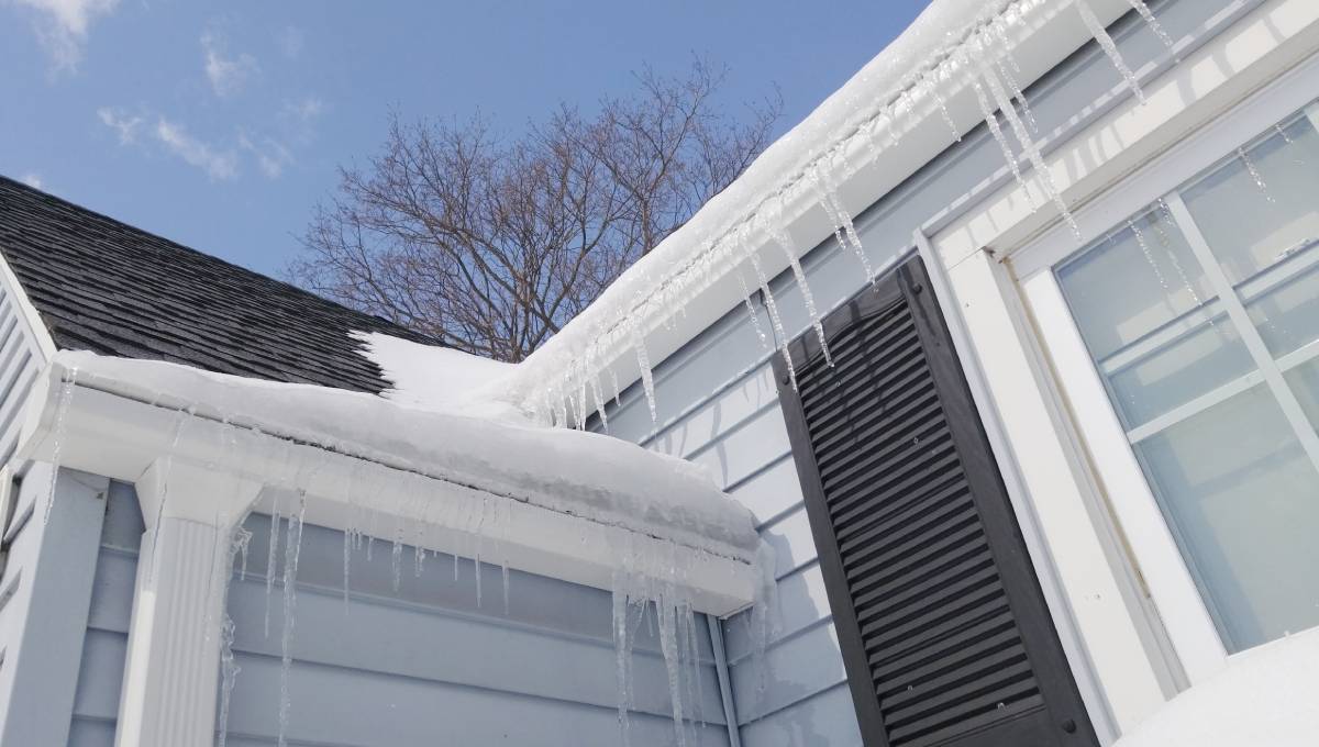 A cozy gray home in the winter season. Snow is on the roof and several icicles are hanging from the windows and gutters.