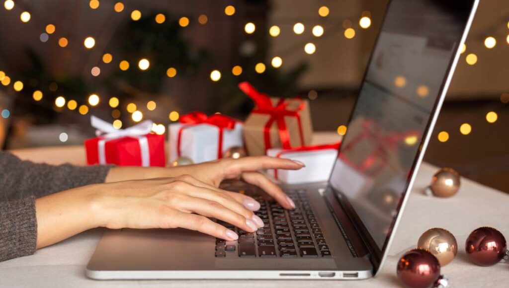 A woman types on a laptop while surrounded by festive gifts, Christmas decorations, and blurred holiday lights.