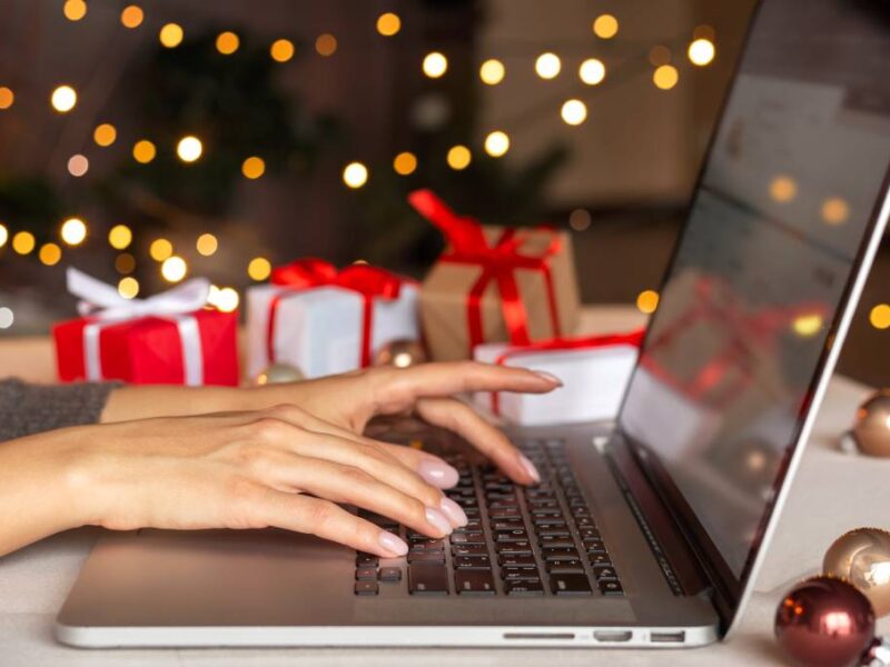 A woman types on a laptop while surrounded by festive gifts, Christmas decorations, and blurred holiday lights.