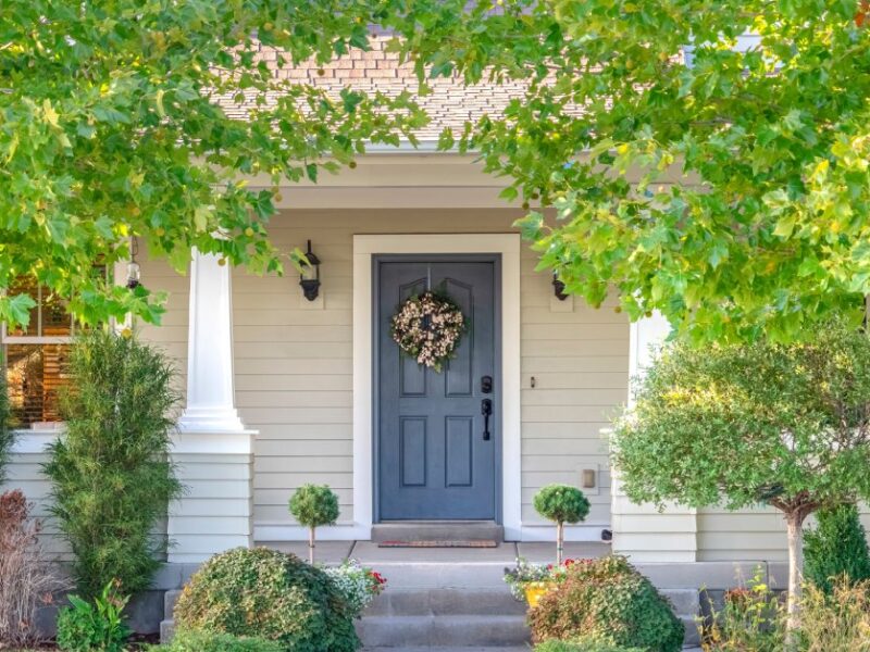 A blue front door with a wreath on it. The door is framed by leaves of trees planted on either side of the house.