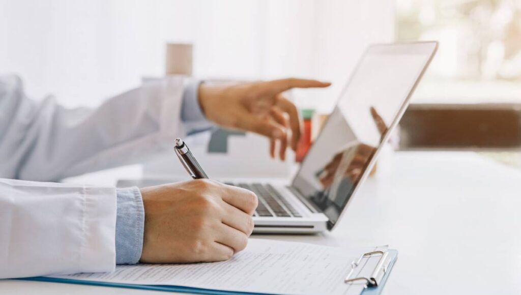 A doctor in a hospital setting writing with a pen on a clipboard while reviewing information from their laptop.