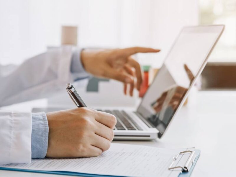 A doctor in a hospital setting writing with a pen on a clipboard while reviewing information from their laptop.