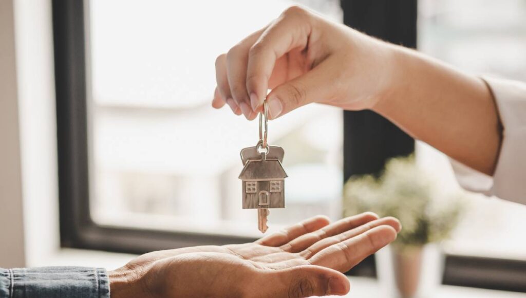 A person's hand dangles a key ring with a key and a house charm on it as another hand reaches out to receive it.