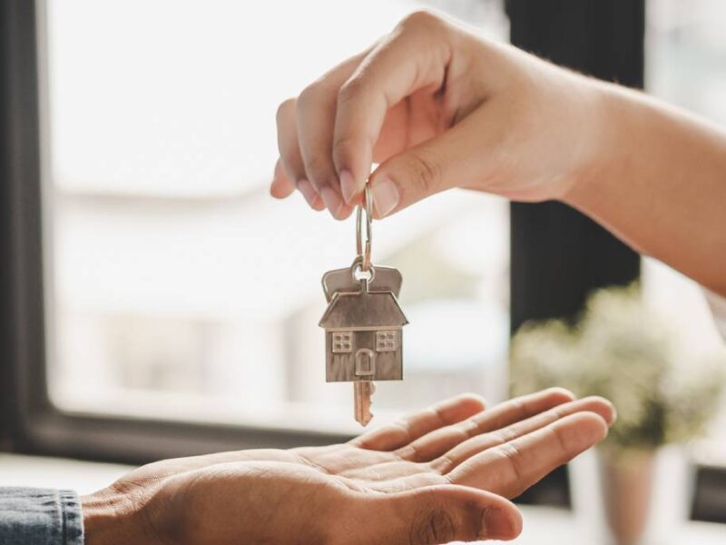 A person's hand dangles a key ring with a key and a house charm on it as another hand reaches out to receive it.