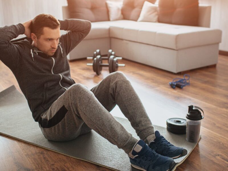 A man wearing a gray jacket and sweatpants sitting on a yoga mat in his home next to a set of dumbbells and a water bottle.