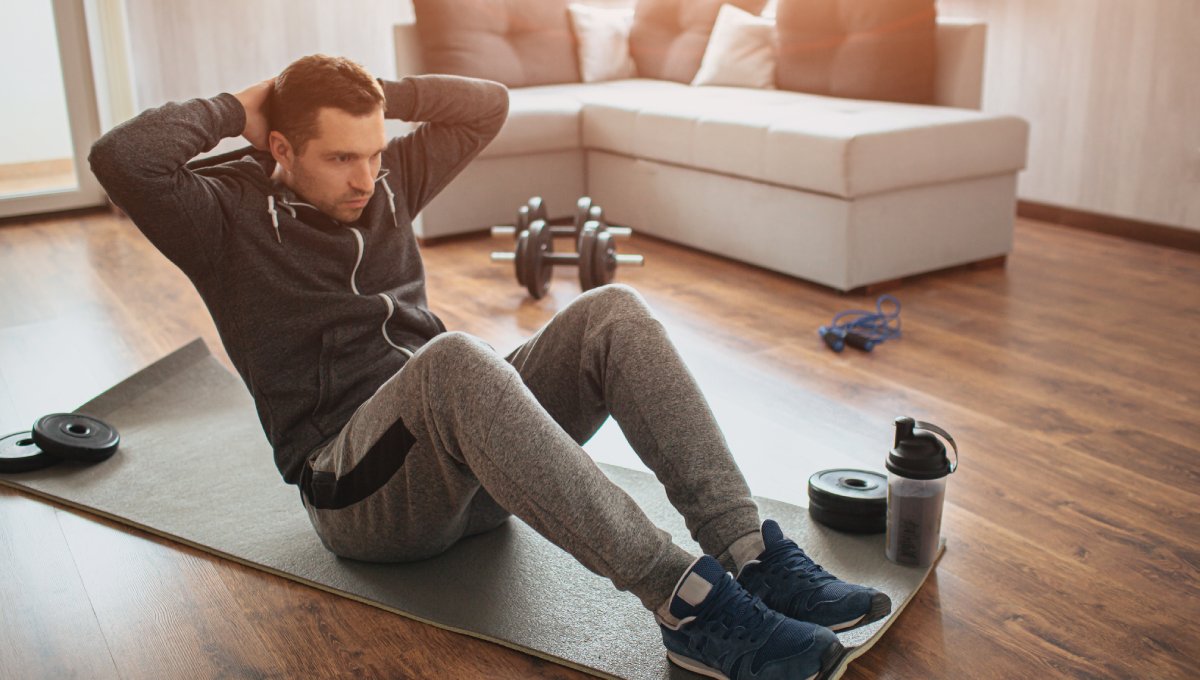 A man wearing a gray jacket and sweatpants sitting on a yoga mat in his home next to a set of dumbbells and a water bottle.